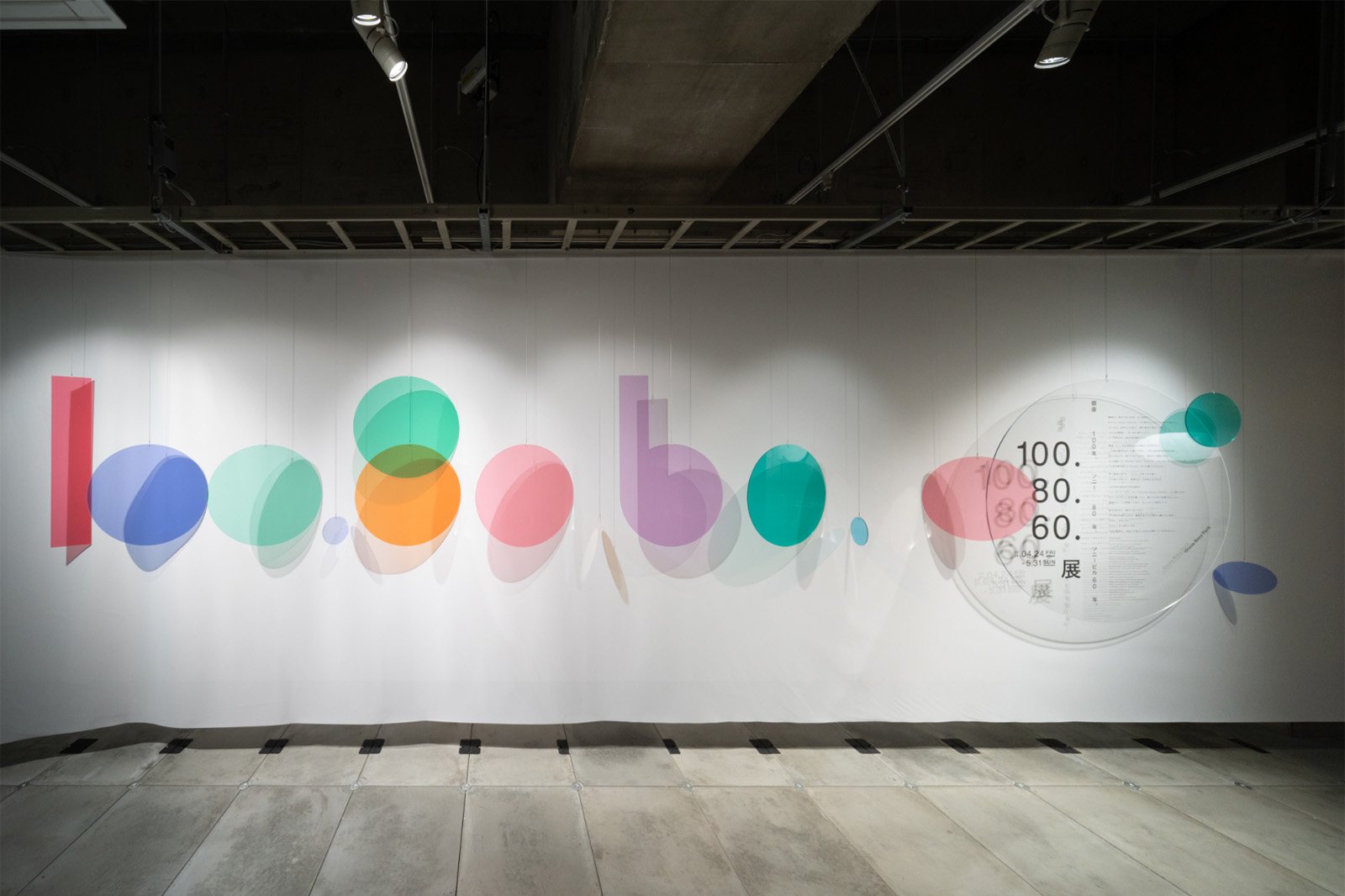 展示導入のビジュアル。壁面に色とりどりの円形グラフィックが並び、「100.80.60.展」のロゴとともに本展のコンセプトを象徴的に示している。